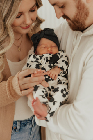A young couple holds their newborn baby in an indoor studio wearing  neutral color matching outfits. Photography by Hannah Lylene Photography.