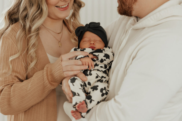 A young couple holds their newborn baby in an indoor studio wearing  neutral color matching outfits. Photography by Hannah Lylene Photography.