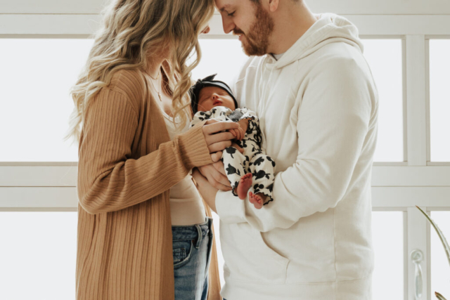 A young couple holds their newborn baby in an indoor studio wearing neutral color matching outfits. Photography by Dallas Photographer Hannah Lylene Photography