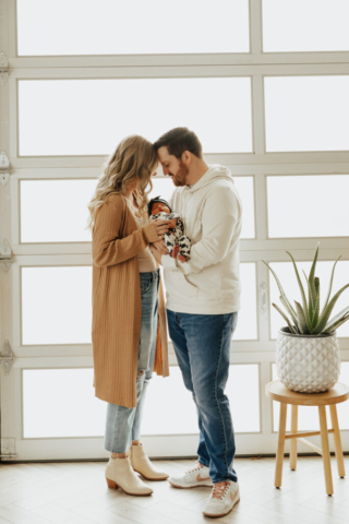 A young couple holds their newborn baby in an indoor studio wearing  neutral color matching outfits. Photography by Hannah Lylene Photography.