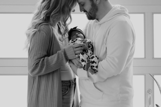 A young couple holds their newborn baby in an indoor studio wearing neutral color matching outfits. Photography by Dallas Photographer Hannah Lylene Photography