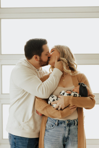 A young couple holds their newborn baby in an indoor studio wearing  neutral color matching outfits. Photography by Hannah Lylene Photography.