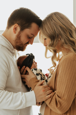 A young couple holds their newborn baby in an indoor studio wearing  neutral color matching outfits. Photography by Hannah Lylene Photography.