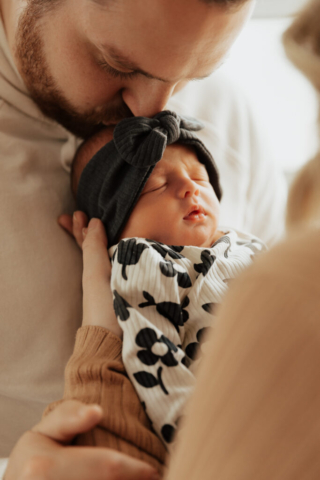 A young couple holds their newborn baby in an indoor studio wearing  neutral color matching outfits. Photography by Hannah Lylene Photography.