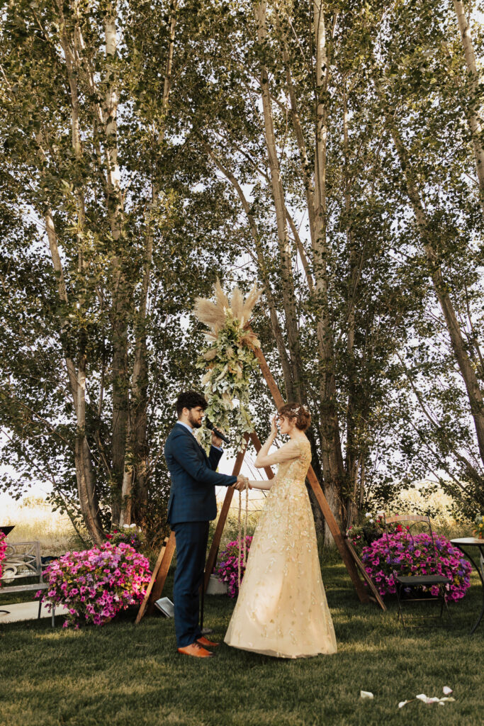A bride in a golden dress listens to her groom's vows at their backyard wedding ceremony