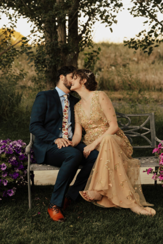 A bride in a gold dress and her groom in a blue suit sit on a wedding bench