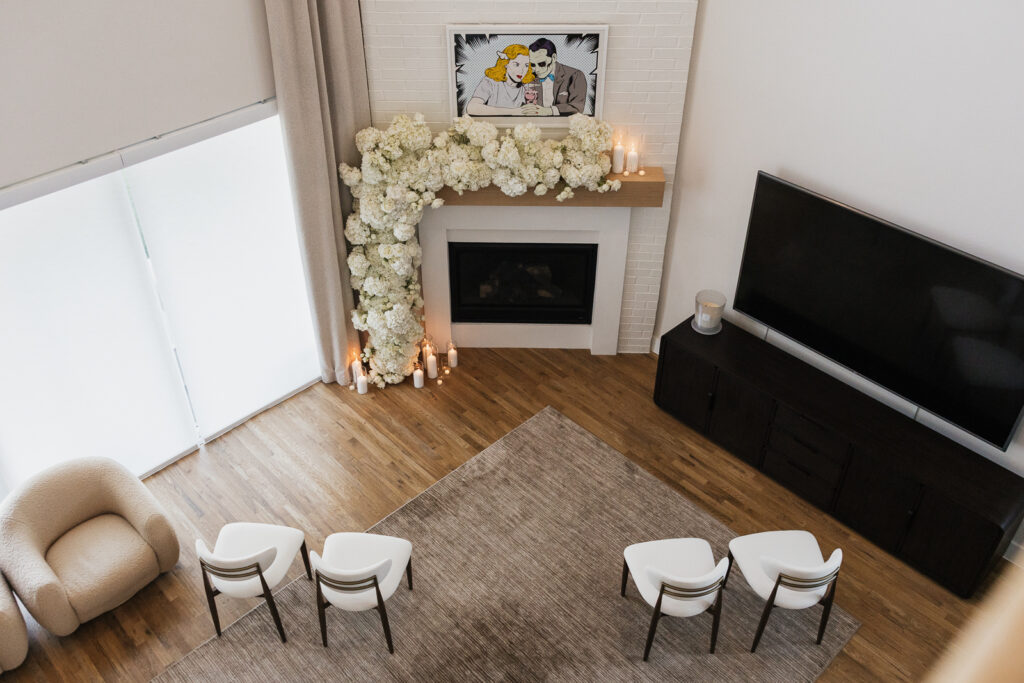 An aerial view of a modern aesthetic home and it&rsquo;s living room area. A white floral arch lines, the fireplace with modern art above it. There are four chairs for a wedding guests. Timeless indoor wedding in Dallas, captured with a refined, editorial approach by DFW documentary wedding photographer.