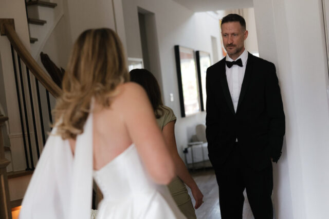 A groom looks affectionately at his bride as she walks down the stairs during their private first look. Timeless indoor wedding in Dallas, captured with a refined, editorial approach by DFW documentary wedding photographer.