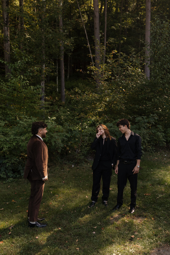 Three people standing in a wooded area, two in black suits, one in brown.