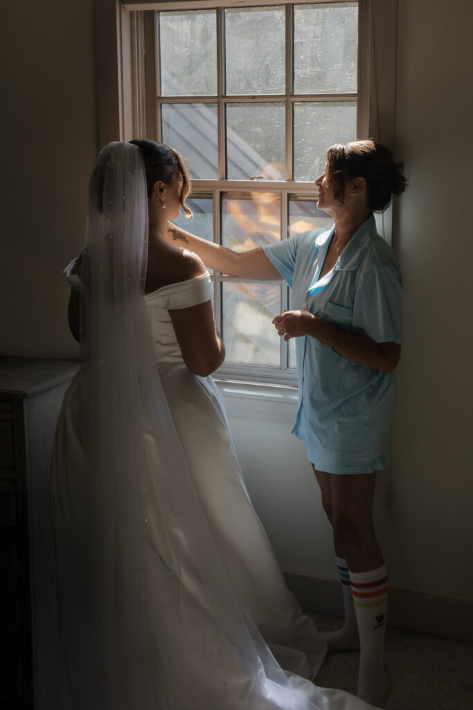 Bride in wedding dress with veil being helped by a woman in a blue robe near a window.