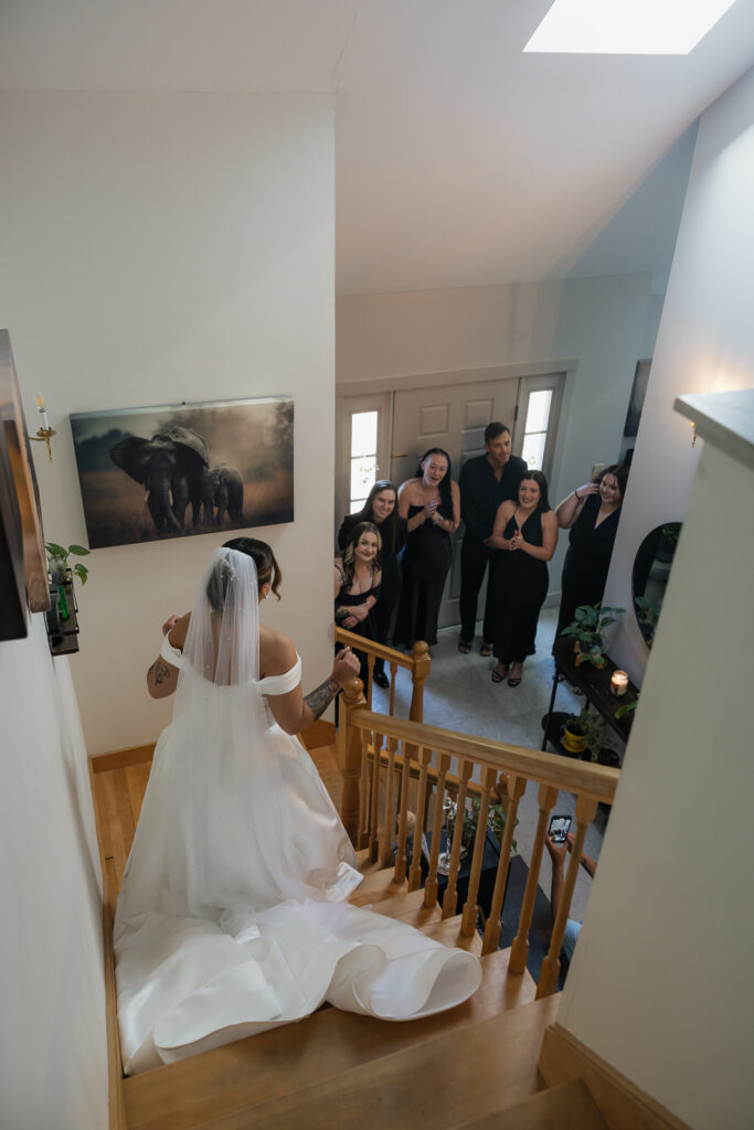 Bride in white wedding dress walking down staircase with bridesmaids clapping
