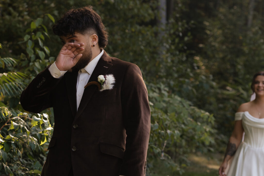 Groom wipes tears as bride approaches in forest setting