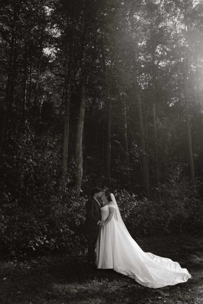 Black and white photo of a bride and groom kissing in a forest, surrounded by tall trees.
