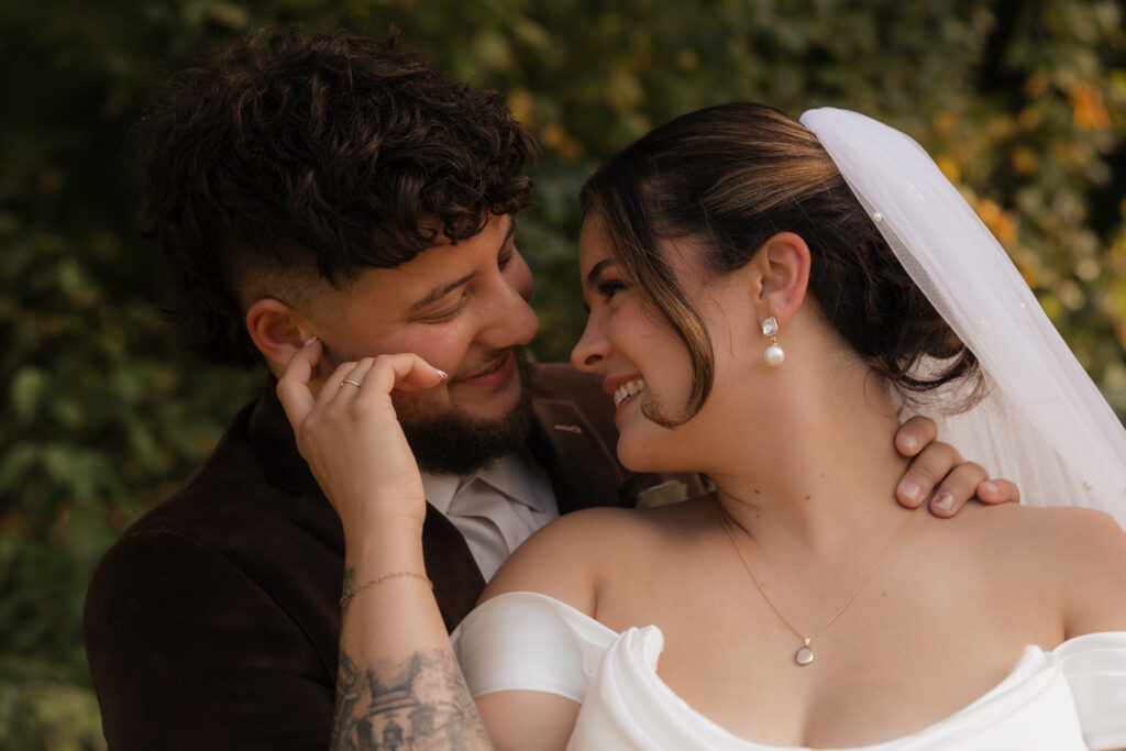 Close-up of a happy newlywed couple, the bride in a white gown and veil, groom in a brown suit, smiling at each other.