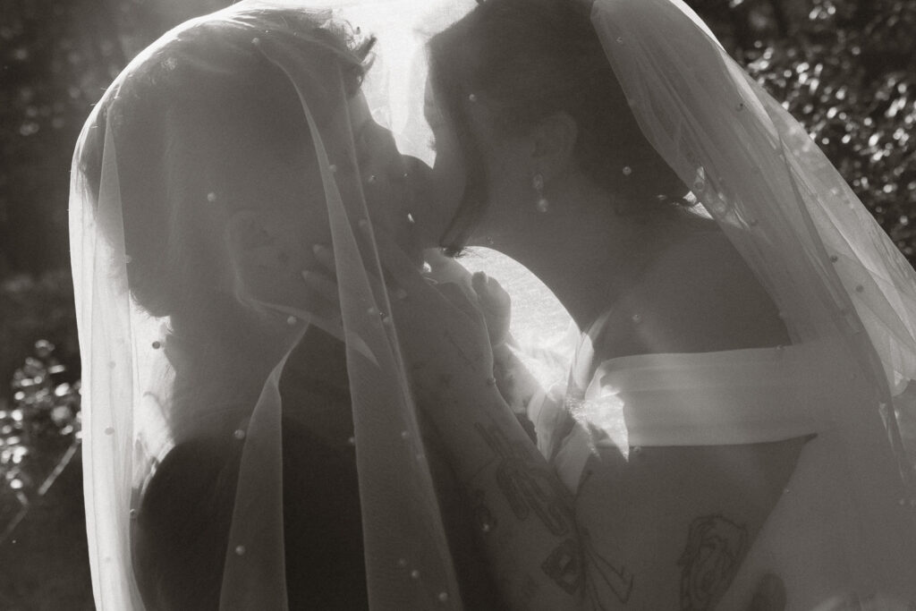Black and white photo of a couple kissing beneath a veil during their wedding ceremony