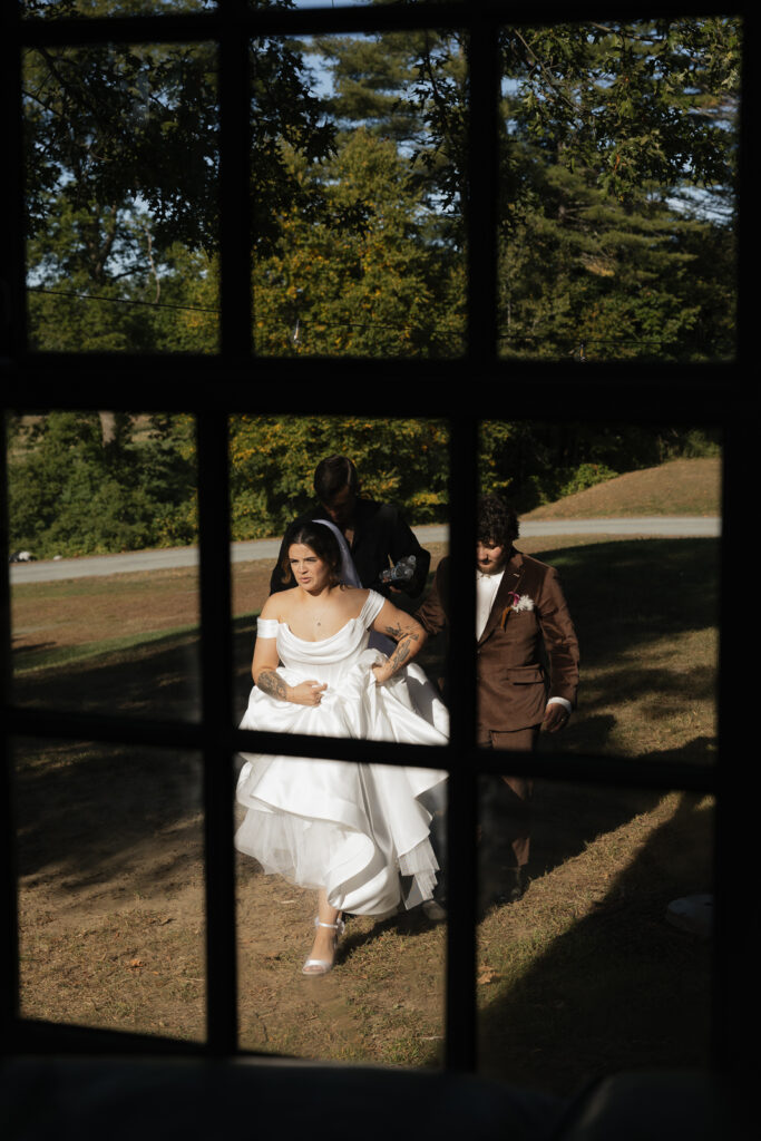 Bride in white dress and groom in brown suit seen through window pane outdoors.
