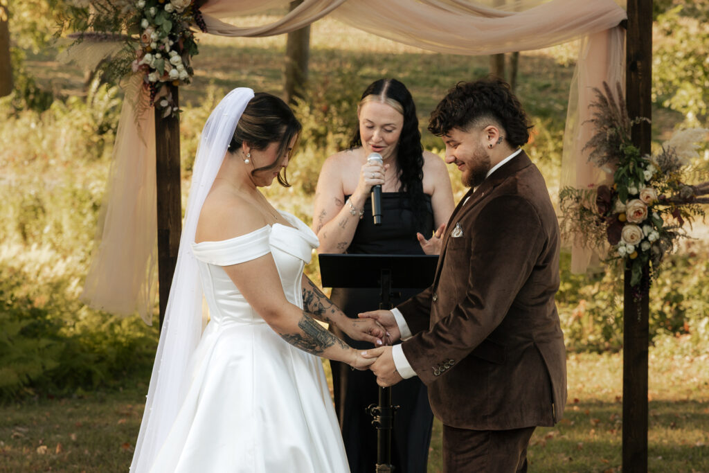 Wedding ceremony outdoors with bride in white gown and groom in brown suit holding hands under floral arch
