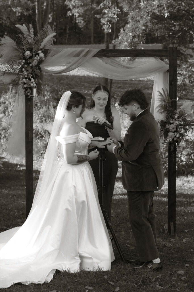 Bride and groom exchange rings during an outdoor wedding ceremony under an arch adorned with flowers and fabric.