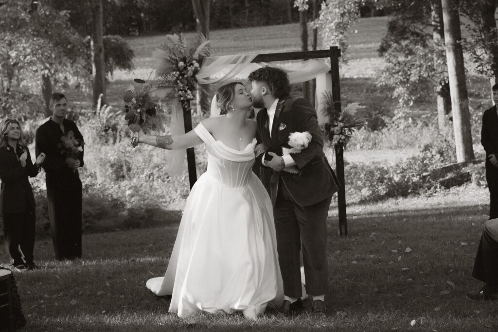 Bride in white off-the-shoulder gown kissing groom in suit outdoors