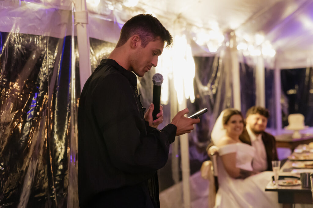 Man giving a speech at a wedding reception with bride and groom smiling in the background.