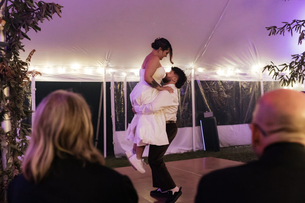 Man lifting woman in white dress on dance floor under a tent with string lights.