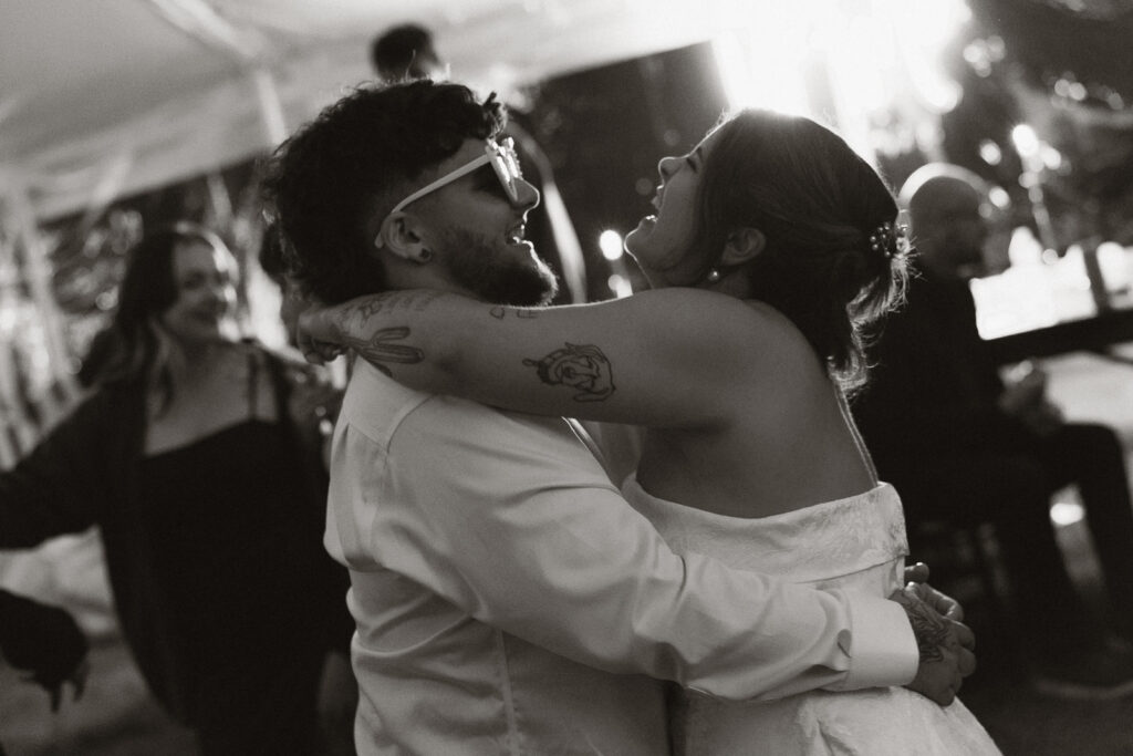 Black and white photo of a couple dancing joyfully at a wedding reception, with others in the background.