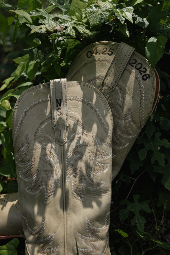 Close-up of beige cowboy boots with intricate stitching, customized with initials "NS" and the date "04.25.2026", nestled in green foliage.