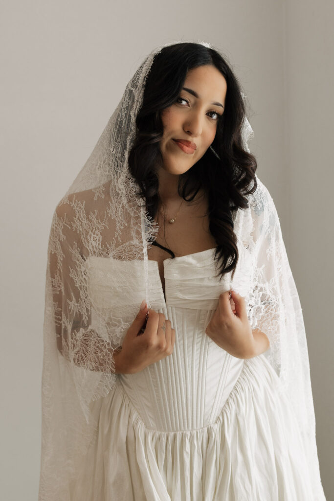 Beautiful bride with dark curly hair wearing a white corset wedding dress and a sheer lace mantilla veil, looking at the camera.