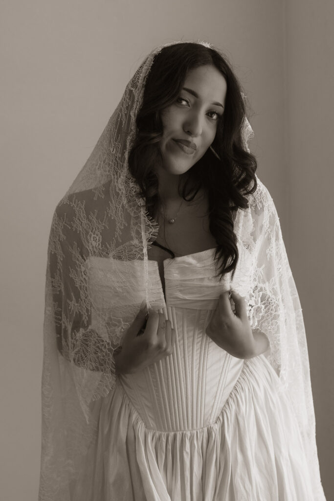 Monochrome portrait of a bride adjusting her white wedding corset and lace veil, looking at the camera.