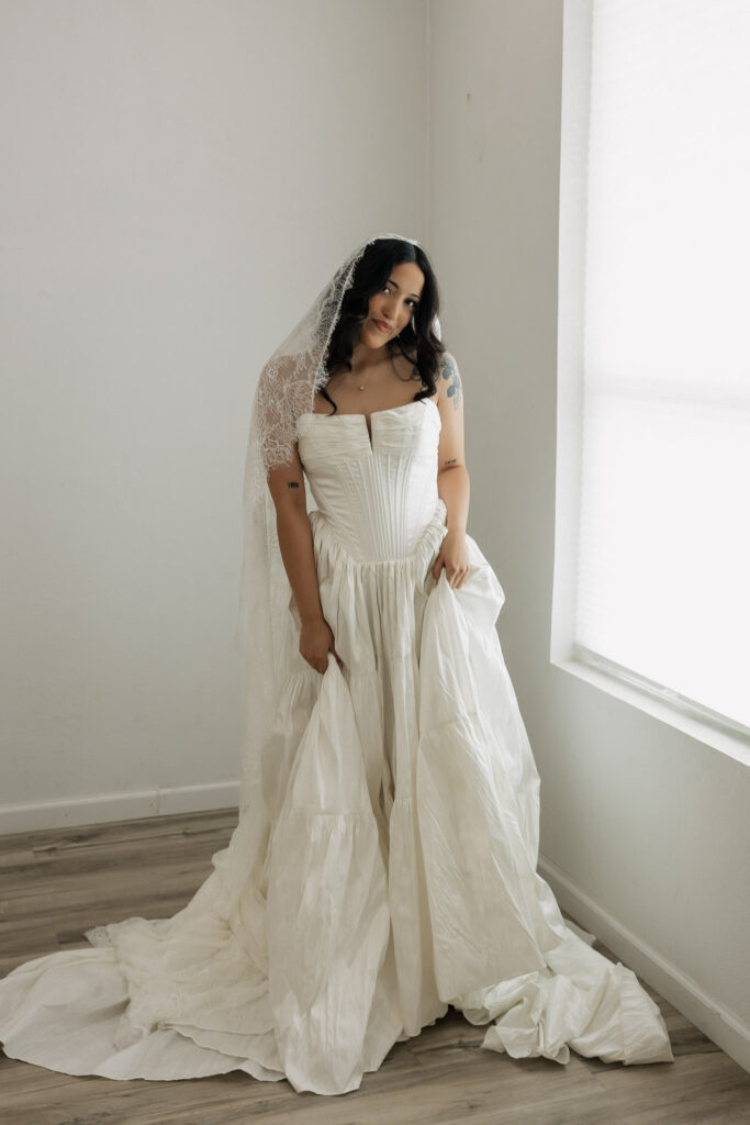 Beautiful bride in a white wedding dress with a lace veil, posing by a window, ideal for wedding fashion.