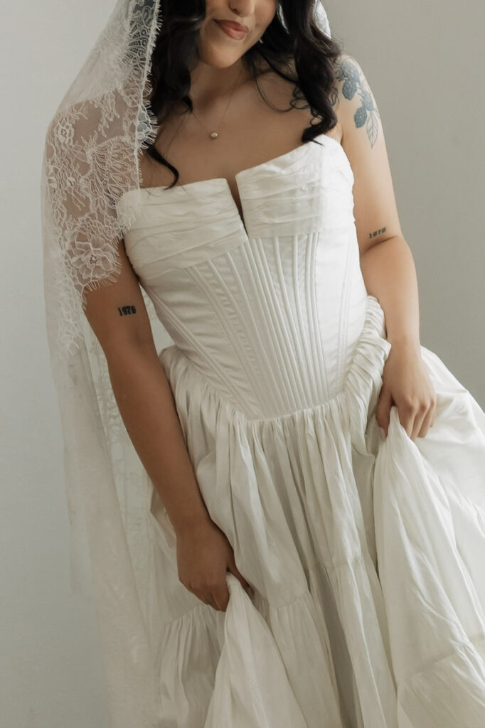 Beautiful bride in a white strapless wedding gown with a corset bodice and tiered skirt, holding up her dress with a delicate lace veil over her shoulder.