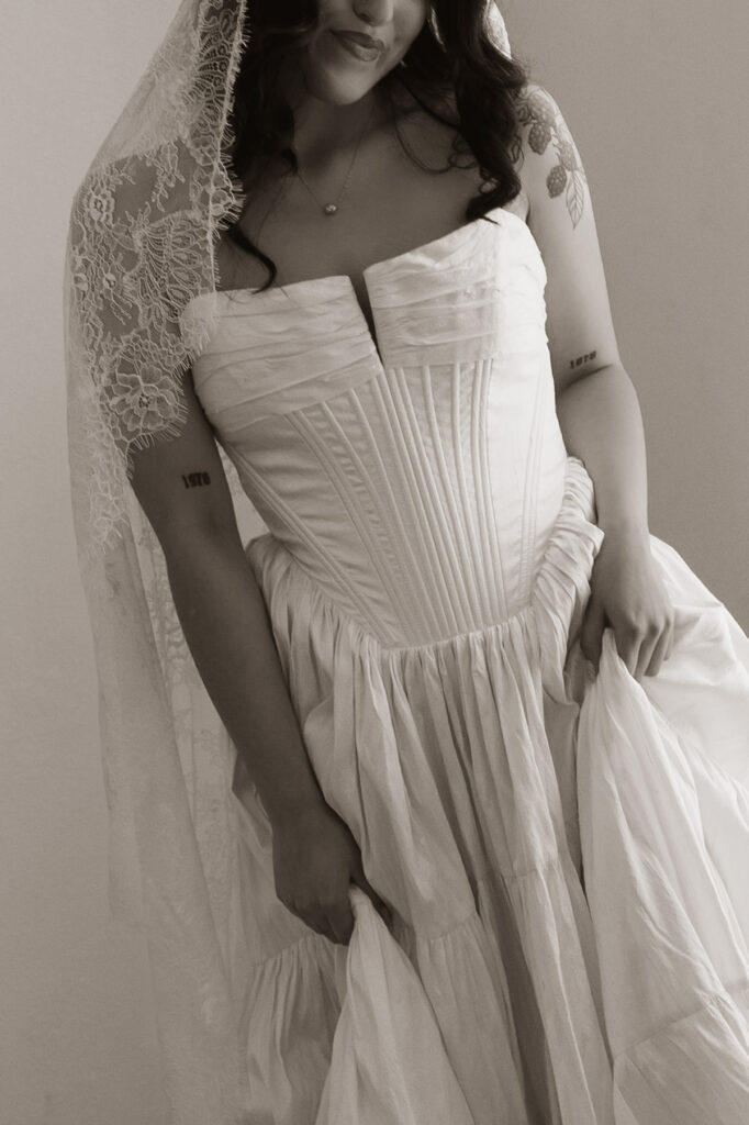 Monochrome close-up of a bride in a strapless white wedding dress with a lace-trimmed veil. She has tattoos on her arms.