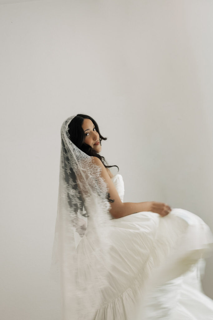 Beautiful bride in a white wedding gown and elegant lace veil twirling, captured in a joyful moment against a minimalist background.
