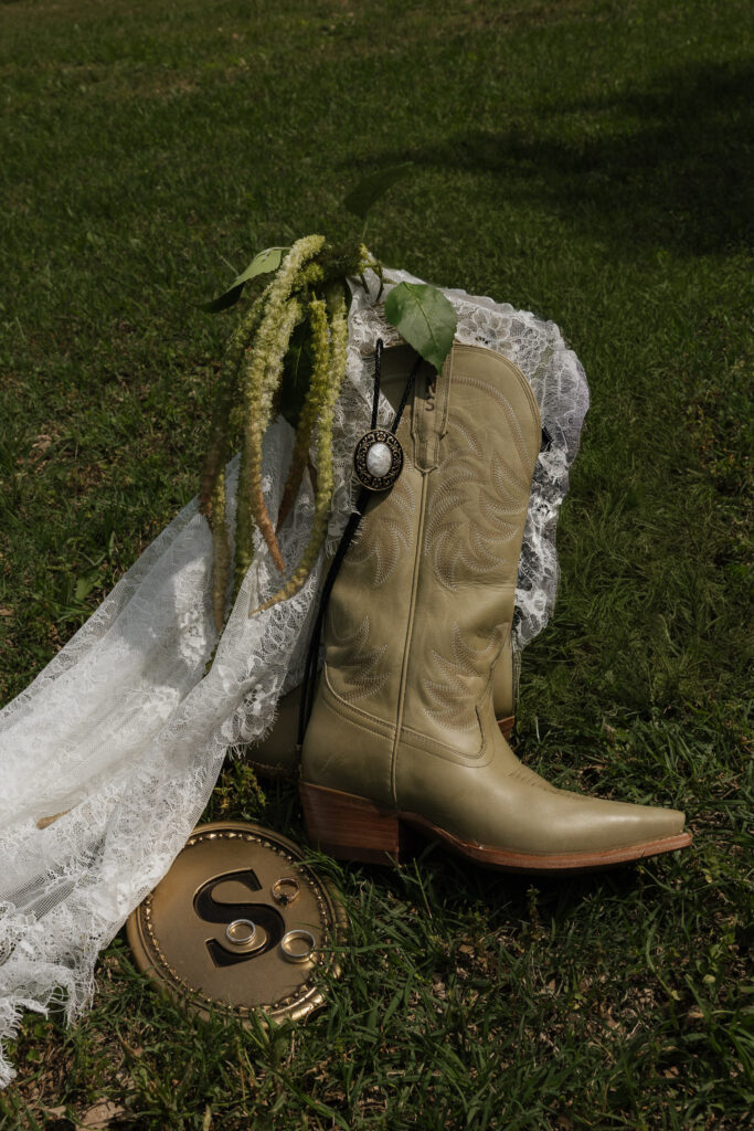 Tan cowboy boots with a bolo tie and lace veil on grass, next to a golden S initial with wedding rings.