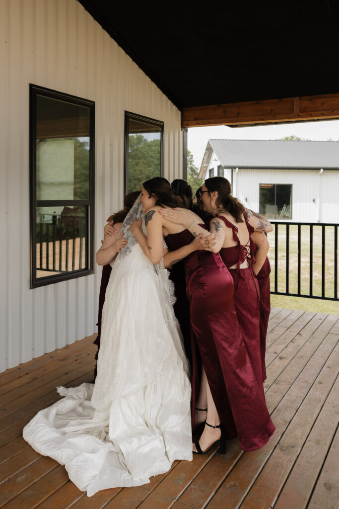 A bride in a white gown and veil hugs her four bridesmaids in dark red dresses on a wooden porch outside a modern white building.