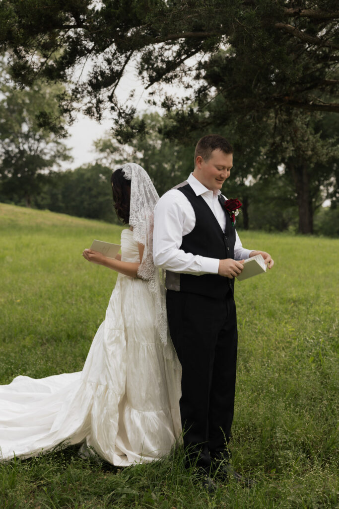 Bride and groom standing back-to-back in a field, reading private vows before their wedding ceremony.