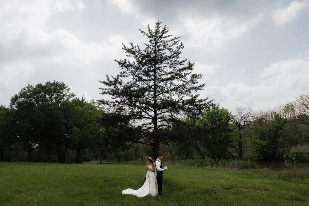 Bride and groom standing by a large tree in a field, exchanging vows under a cloudy sky.