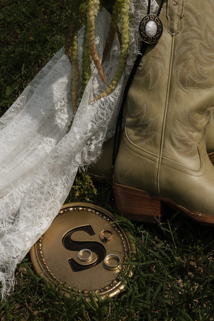 Close-up of a rustic wedding arrangement featuring a cowboy boot, bolo tie, lace fabric, a bouquet, and wedding rings on a golden "S" plate amidst green grass, ideal for themed ceremonies.