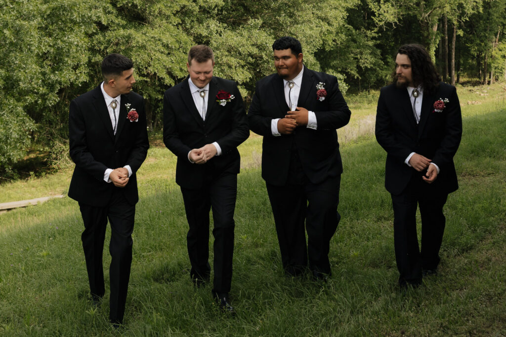 Four groomsmen in black suits with bolo ties and burgundy boutonnieres walk through green grass, celebrating a wedding day.