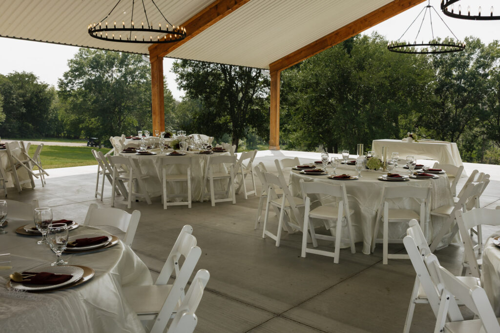 An outdoor wedding reception setup with round tables, white chairs, and elegant place settings under a covered pavilion surrounded by green trees.