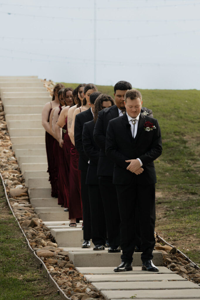 Groomsmen in black suits and bridesmaids in maroon dresses stand in a line on outdoor steps at a wedding ceremony.