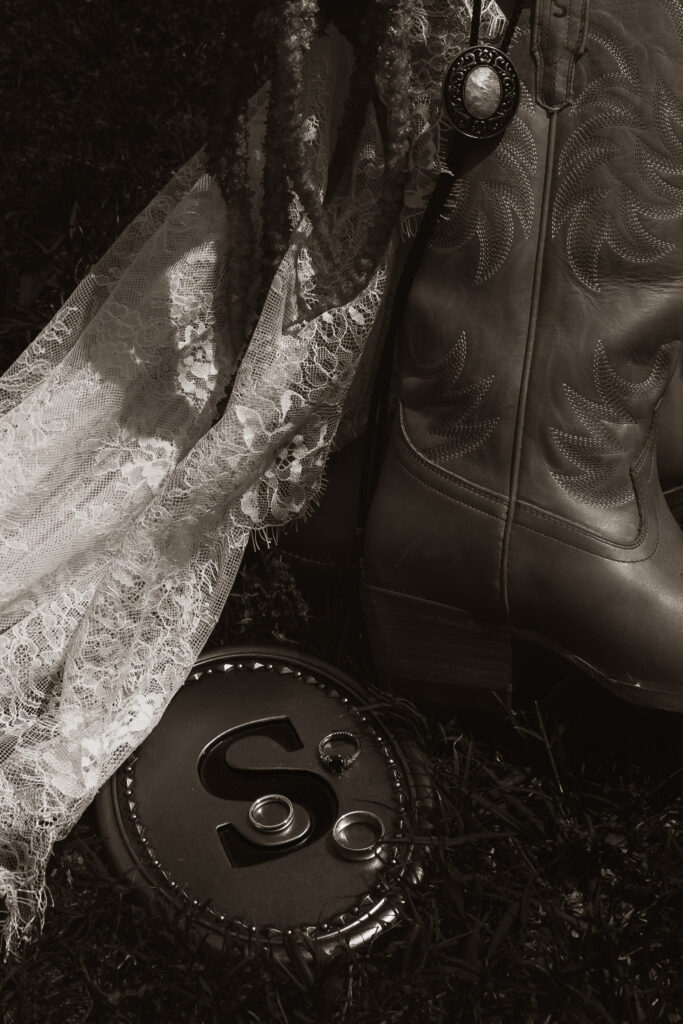 Monochrome image featuring a white lace wedding dress, brown cowboy boots with intricate stitching, and wedding rings.