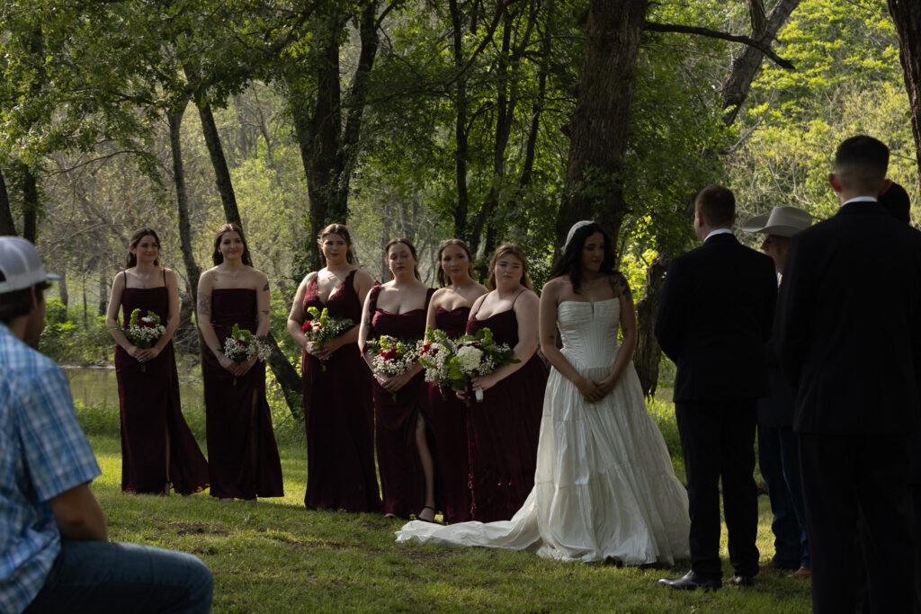 Outdoor wedding ceremony with bridesmaids and bride in white dress, groomsmen in suits.