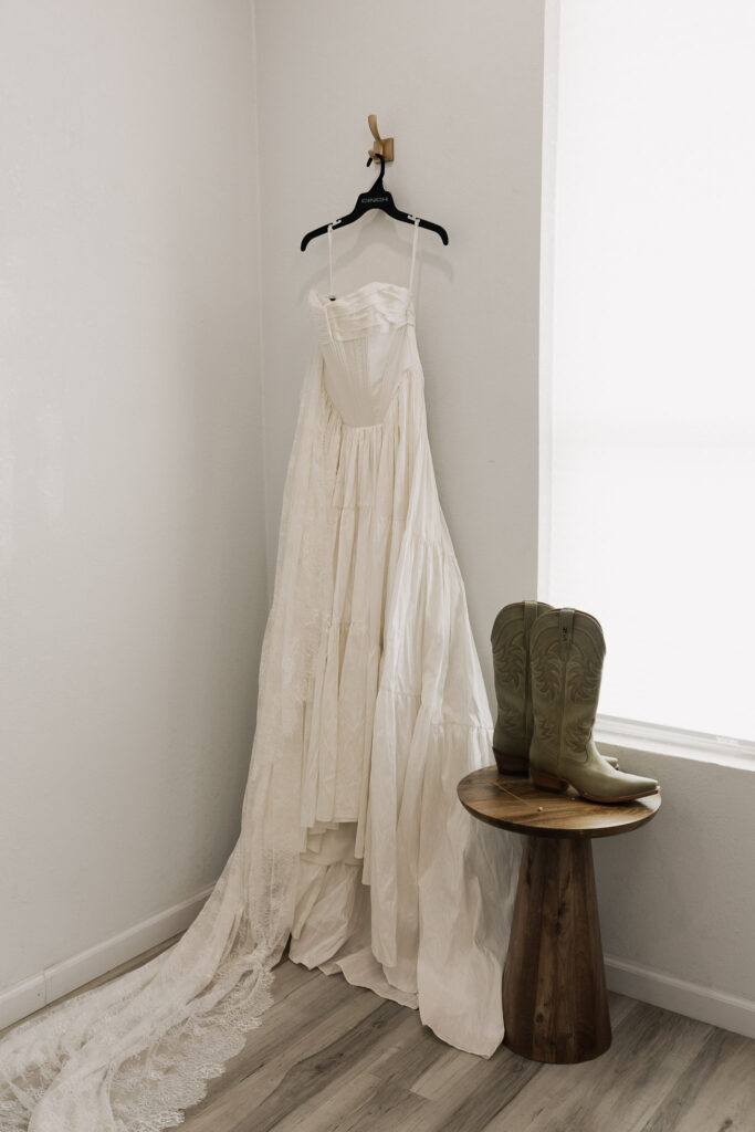 An ivory tiered wedding dress with a long lace train hangs on a wall next to a wooden pedestal table with light green cowboy boots.