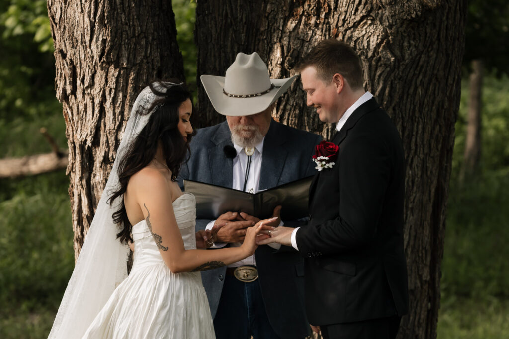 Bride and groom exchange rings at outdoor wedding ceremony with officiant in cowboy hat.