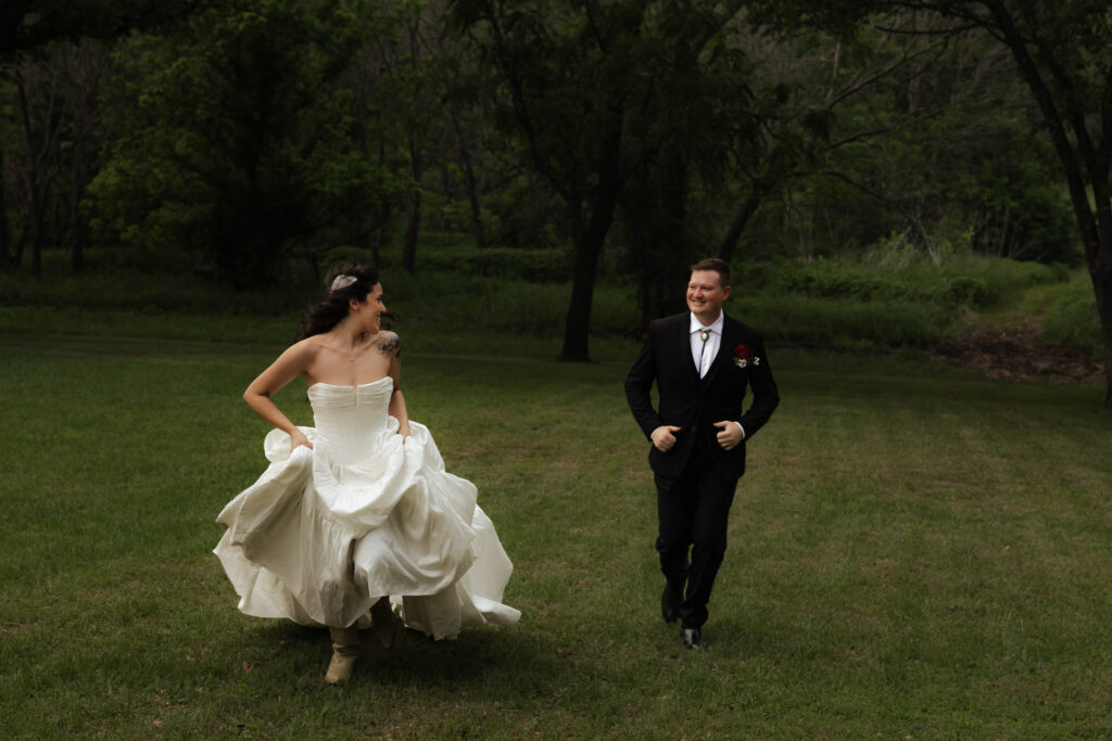 Bride in white dress and groom in black suit running joyfully on a grassy field with trees in the background.