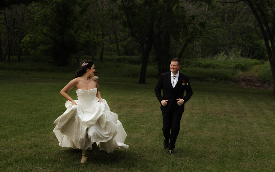 Bride in white dress and groom in black suit running joyfully on a grassy field with trees in the background.