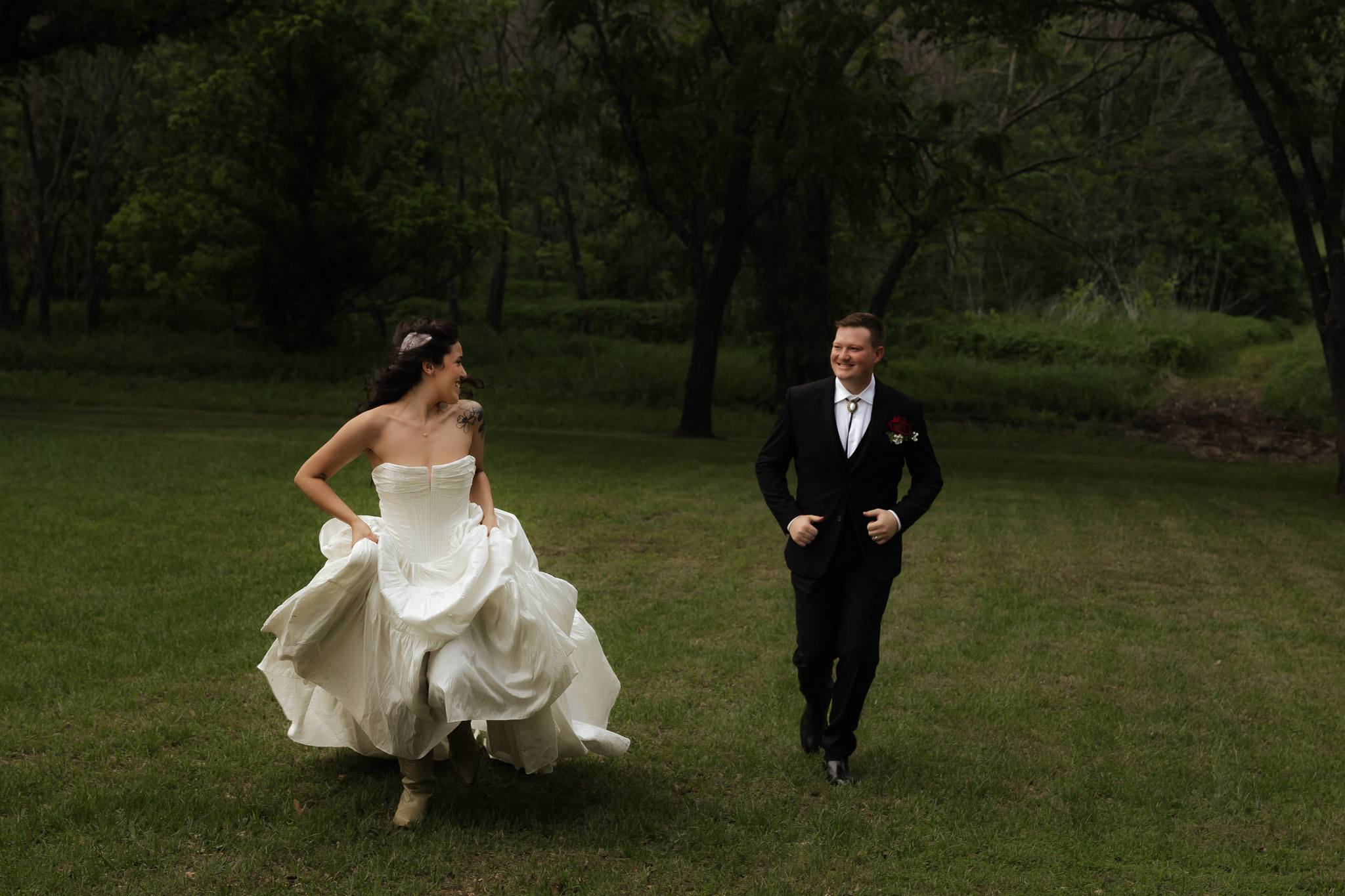 Bride in white dress and groom in black suit running joyfully on a grassy field with trees in the background.