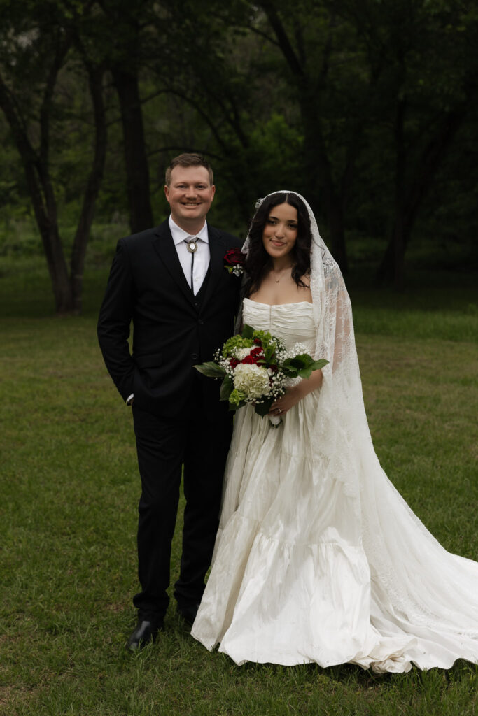 Bride and groom standing outdoors in a park, bride in a white lace gown, groom in a black suit.