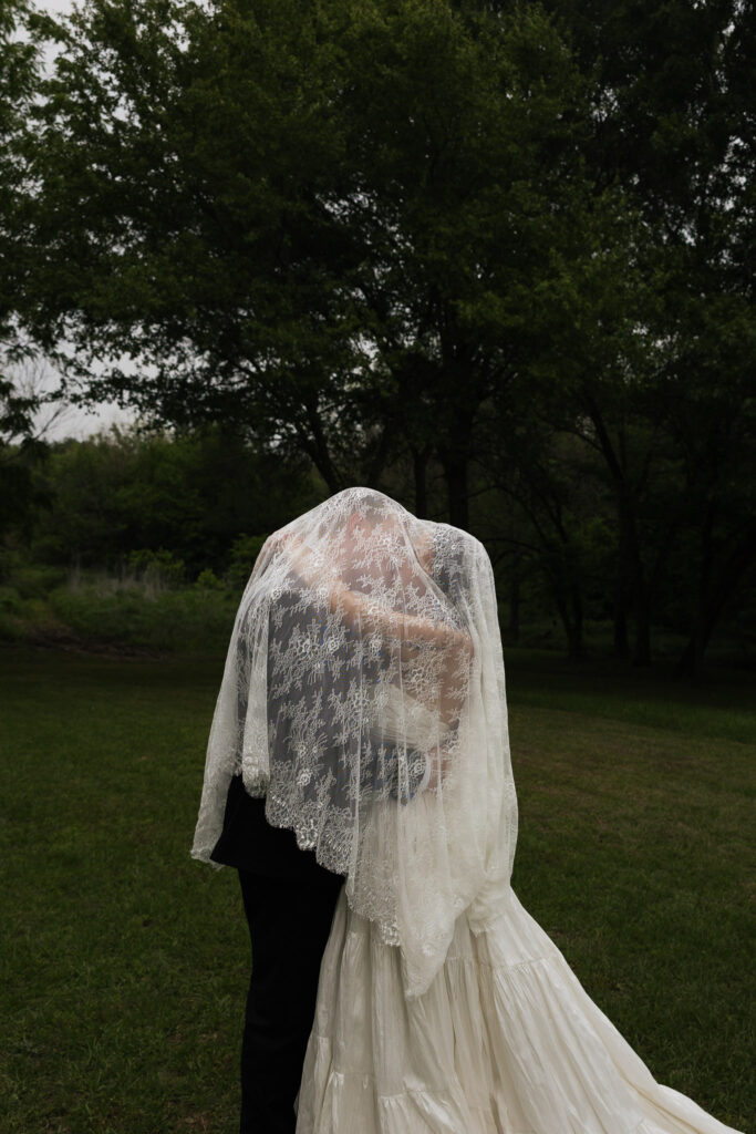 Bride and groom kiss under lace veil outdoors.